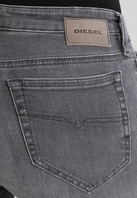 Jeans de mezclilla gris con un bolsillo trasero que presenta un patrón de costura distintivo y un parche de cuero etiquetado como "DIESEL" en la cintura.