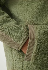 Blød grøn fleecejakke med tekstureret stof, ribstrikkede olivengrønne detaljer ved ærmer og lomme, med en rummelig håndlomme på siden.