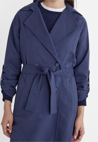 Marineblauwe trenchcoat met een dubbelgeknoopte ontwerp, brede revers, strikceintuur en opgerolde mouwen, gemaakt van een soepele stof.