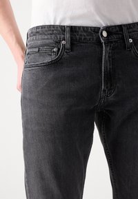 Črne jeanske hlače slim fit, ki imajo klasično zasnovo s petimi žepi, neopaznim šivanjem in srebrnim gumbom za zapiranje.