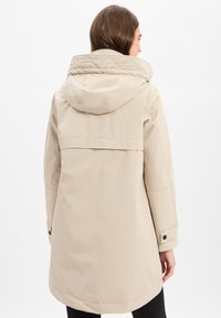 Beige wasserfeste Jacke mit Kapuze, ausgestattet mit einem gesteppte Kragen, Druckknöpfen an den Ärmel und einem leicht verlängerten Rücken für mehr Schutz.
