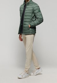 Zelená prošívaná bunda s vysokým límcem, bočními kapsami a úzkým střihem, nošená přes tmavý top, v kombinaci s béžovými kalhotami a bílými teniskami.