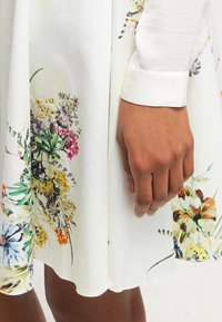 Robe blanche avec des motifs floraux aux couleurs vives. Présente une longue manche avec un poignet. Tissu doux et lisse, au design légèrement fluide.