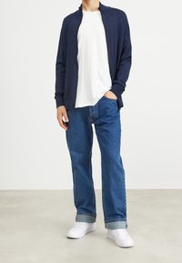 Man som bär en marinblå zip-up cardigan, vit t-shirt, uppvikta blå jeans och vita sneakers, står med en hand i fickan.