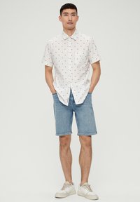 Kurzärmeliges weißes Hemd mit kleinem roten Punktmuster, Knopfverschluss und Tasche, kombiniert mit blauen Jeansshorts und weißen Sneakers.