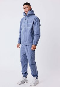 Jonge man in een blauwe hooded trainingsoutfit met ritszakken en witte sneakers, staand tegen een effen lichte achtergrond.