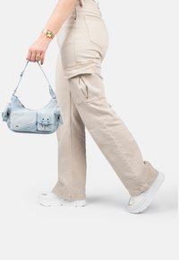 Hellblaues Handtasche mit mehreren Reißverschlusstaschen, getragen von einer Person in beigen Cargo-Hosen und weißen klobigen Sneakern. Glatte Textur.