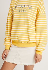 Sudadera a rayas amarillas y blancas con el texto "VENICE VIBES" combinada con una falda mini de mezclilla blanca con bordes deshilachados, en una persona de pie.