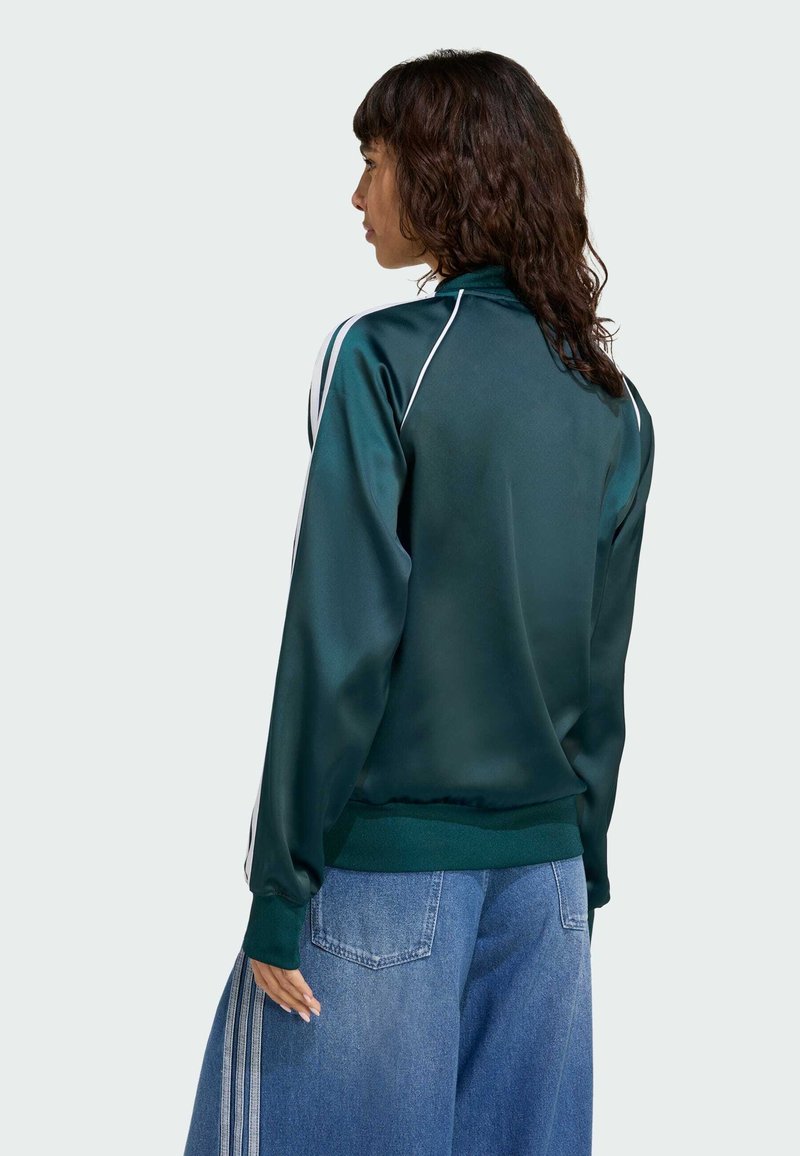 Grøn satinjakke med hvide sidelister, høj krave, ribstrikkede ærmer og kant. Kombineret med brede denim bukser. Set bagfra.