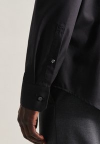 Chemise noire à manches longues avec un poignet boutonné et un tissu texturé. La manche est décontractée, mettant en valeur des détails de couture subtils près du poignet.