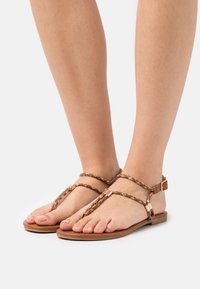 Sandales brunes avec des brides tressées, semelle plate et accents dorés. Design à bout ouvert, adapté au port décontracté.