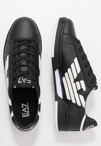 Baskets en cuir noir avec des accents blancs et un design d'aile sur le côté, dotées d'un laçage à l'avant et d'une semelle en caoutchouc texturée.