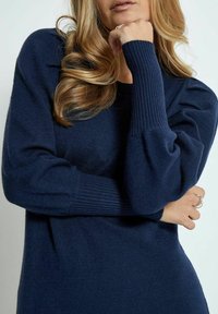 Pull en tricot bleu marine avec des poignets côtelés et des manches bouffantes, doté d'une texture douce et d'une coupe décontractée. Mains reposant sur le bras.