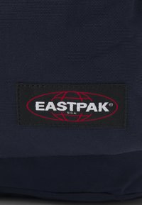 Marinblå tygryggsäck med en svart etikett med röd och vit text "EASTPAK" och en globdesign. Slät yta, robust konstruktion.