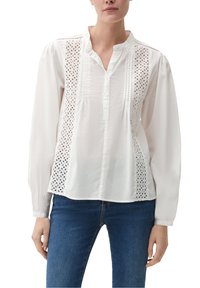 Donna in una camicia bianca a maniche lunghe con pannelli di pizzo e dettagli a cuciture sottili, abbinata a jeans blu, in piedi contro uno sfondo semplice.