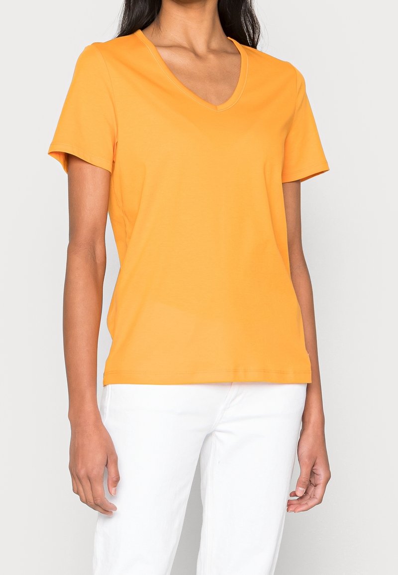 Femme portant un t-shirt orange à manches courtes et col en V, ainsi qu'un pantalon blanc, sur un fond neutre.