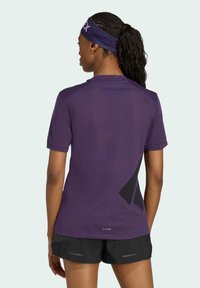 Camiseta atlética morada de manga corta, con textura de malla y diseño de acento negro en la parte posterior, combinada con pantalones cortos negros y una diadema a juego.
