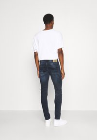 Jeans de mezclilla azul oscuro con un ajuste slim, que presentan una sutil textura desgastada y bolsillos traseros. Combinados con una camiseta blanca sencilla y zapatillas blancas.