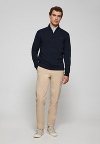 Sudadera con cremallera en color marino con un diseño ajustado, puños acanalados y un pequeño logo rojo. Combinada con chinos beiges y zapatillas claras.