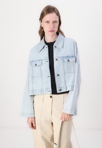 Giacca di jeans corta di colore blu chiaro con due tasche sul petto, chiusura a bottoni e una vestibilità rilassata. Indossata sopra una maglietta nera e abbinata a pantaloni crema.