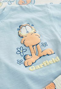Lyseblå t-shirt med et tegneseriekatte-design. Katten er orange med store øjne, omkranset af broderede blomster, med "Garfield" i hvid tekst.