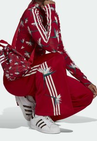 Red tracksuit with floral pattern, white stripes on sleeves and legs, paired with white sneakers featuring black and gold accents, and a matching bag.