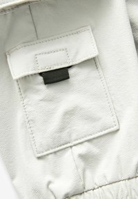 Tissu gris clair avec une texture lisse, doté d'une poche latérale, maintenue par une sangle noire, et avec des coutures froncées en bas.