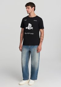 T-shirt nera in cotone con un grande logo PlayStation bianco, accompagnato da testo in giapponese; abbinata a jeans di denim azzurro chiaro e sneakers bianche.