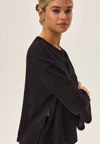Czarna bluzka z długim rękawem o teksturowanym, dzianinowym wzorze, z bocznymi rozcięciami i okrągłym dekoltem. Materiał wydaje się lekki i przewiewny.