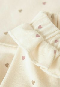 Licht crèmekleurige stof met kleine rozen en bruine hartjespatronen. Heeft een gerimpelde halslijn en een geribbelde textuur, wat een zachte uitstraling geeft.