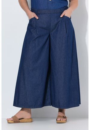 Persona che indossa pantaloni in denim blu scuro a gamba larga con vita elastica e mani nelle tasche, abbinati a sandali con zeppa in legno color marrone chiaro.