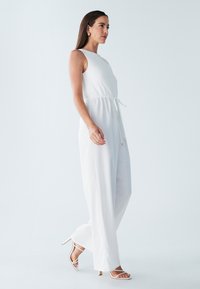 Witte jumpsuit met gestructureerde top, trekkoord in de taille en brede pijpen. Draagt met witte hakken met bandjes. Eenvoudig, elegant ontwerp.
