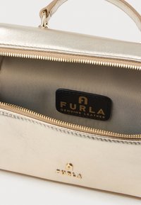 Zelta tonēta ādas rokassoma ar gludu tekstūru, ar rāvējslēdzēja atvēršanu un iekšpusi ar svītrotu rakstu. Iekšpusē ir Furla logo detaļa.