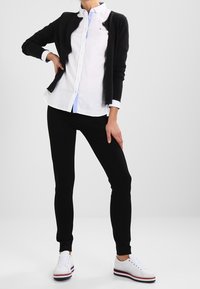 Cárdigan negro sobre una camisa blanca de botones, combinado con pantalones jogger negros y zapatillas blancas con acentos rojos y azules. Diseño simple.