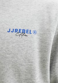 Γκρι ύφασμα με μπλε κείμενο "JJREBEL®" και μαύρο πλάγιο κείμενο "Edition" τυπωμένα πάνω του.