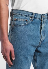 Siniset farkut, joissa on keskikorkea vyötärö, kupariset niitit, etutaskut ja teksturoitu kangas, korostavat klassista rentoa muotoilua.