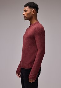 Pull en tricot bordeaux avec un col rond et des poignets côtelés, présentant une finition texturée. La vue de côté montre une coupe décontractée et des manches longues.