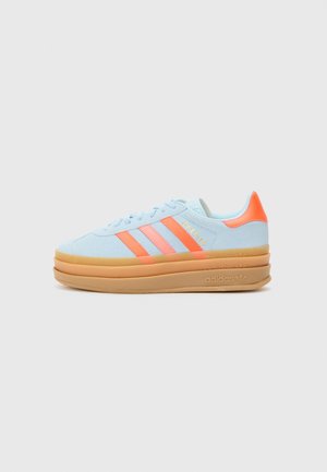 Baskets Adidas bleu clair avec des bandes orange et un languette au talon, semelle plateforme épaisse beige et inscription dorée « Gazelle » sur le côté.