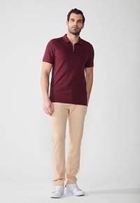 Burgunderfarget poloskjorte med korte ermer og glidelås på kragen, kombinert med lyse beige bukser og hvite sneakers, satt mot en nøytral bakgrunn.