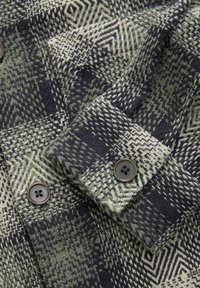 Tissu en laine à motif chevrons noir et gris avec deux boutons noirs ronds cousus sur un revers de manche plié.