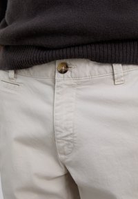 Pantalones beige con cierre de botón, usados con un suéter de canalé marrón oscuro metido ligeramente en la cintura.
