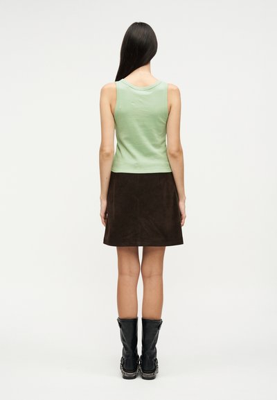 Γυναίκα με μακριά σκούρα μαλλιά, που φοράει ένα αμάνικο ανοιχτοπράσινο τοπ, σκούρα καφέ φούστα και μαύρες μπότες μέχρι τη μέση της γάμπας, στέκεται γυρισμένη με την πλάτη προς το μέρος μας σε λευκό φόντο.