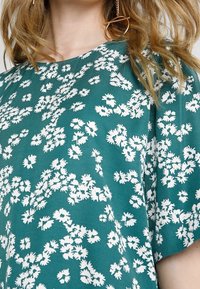 Top de manga corta en color teal con un patrón floral blanco. La tela parece ligera y suave, con un escote redondo y un corte holgado.