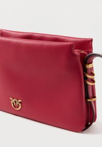 Punane nahast käekott, millel on sile tekstuur. Esiplaanil on nähtav kullatooniline logo ja dekoratiivne riistvara, millel on külgedel rõngad. Lukustatav tõmblukk.