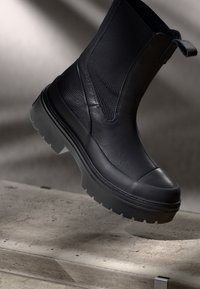 Stivale alla caviglia nero con tomaia in pelle textured, dettagli in rete, punta rotonda e suola in gomma spessa con scanalature profonde.
