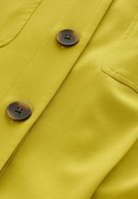 Detailný záber na žltú tkaninu s dvoma hnedými gombíkmi a viditeľným prešitím na odeve.