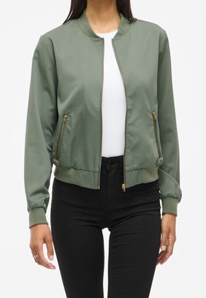 Kobieta ubrana w oliwkowo-zieloną kurtkę bomber z zamkiem błyskawicznym, z prążkowanymi mankietami i dolnym ściągaczem, na białej bluzce i czarnych spodniach, stojąca z rękami w pozycji swobodnej.