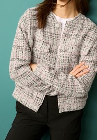 Giacca in tweed a quadri grigia e rosa con scollo rotondo, bottoni a pressione e finitura testurizzata. Indossata sopra una camicia bianca, abbinata a pantaloni neri.