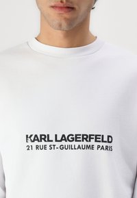 Bijela dukserica s rebrastim okovratnikom i odvažnim crnim slovima: "KARL LAGERFELD 21 RUE ST-GUILLAUME PARIS" na prednjem dijelu.