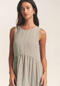 Svetlo siva obleka z okroglim izrezom, brez rokavov, nagubanim pasom in majhnimi temnimi pikami. Mehka, teksturirana tkanina.